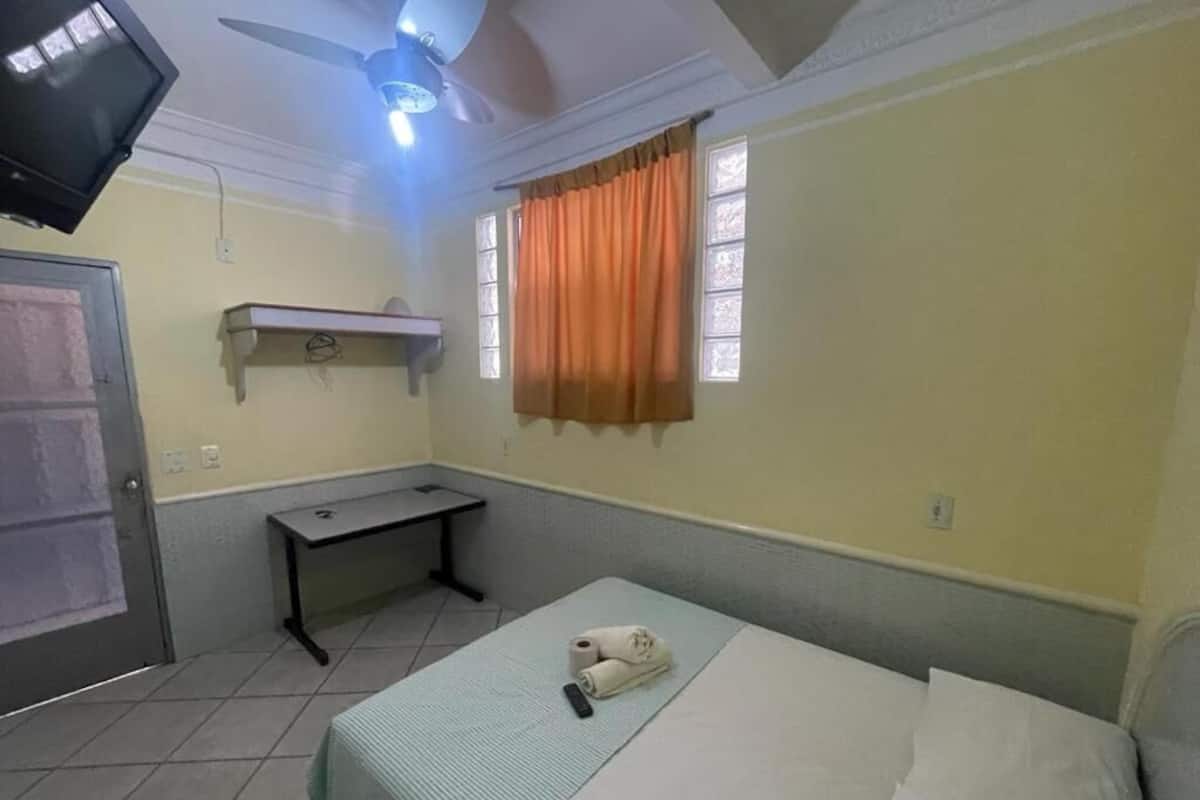 A bedroom with a bed, a desk, a television, and a ceiling fan.