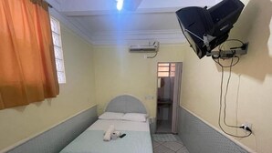 Una habitación de hotel pequeña y estrecha con una cama individual, un ventilador de techo, un equipo de aire acondicionado y un televisor montado en la pared.