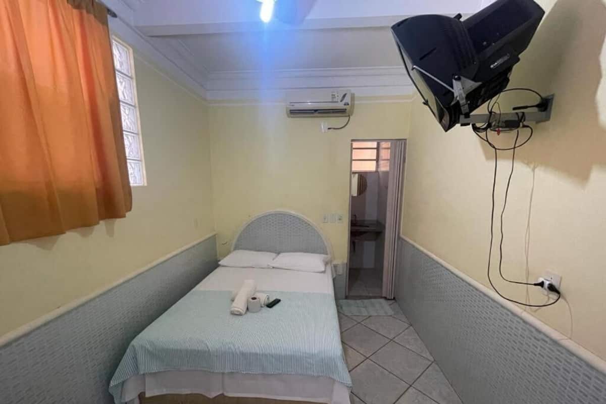 A small, narrow room with a single bed, a ceiling fan, an air conditioning unit, and a wall-mounted projector.