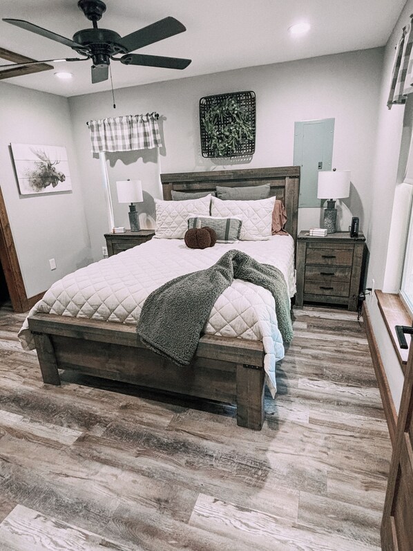 A bedroom with a wooden bed, a ceiling fan, a nightstand with a lamp, and a wall-mounted artwork.