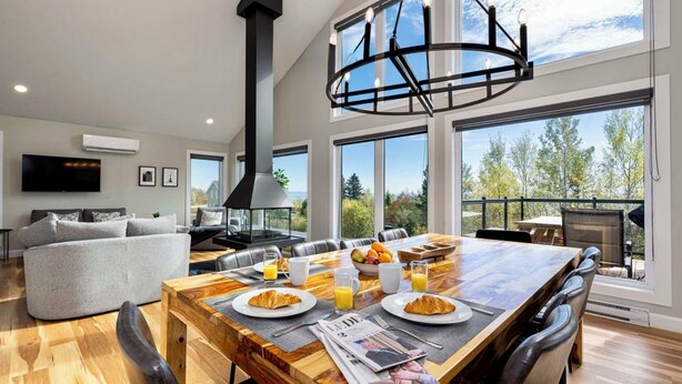 A modern living room with a dining area, large windows, and a fireplace. - Ski & relax with a view of the river - L'Aube Charlevoix (Petite-Rivière-Saint-François)