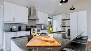 A modern kitchen with white cabinets, a stainless steel range hood, and a large island with a cutting board, wine glasses, and a loaf of bread. - Ski & relax with a view of the river - L'Aube Charlevoix (Petite-Rivière-Saint-François)