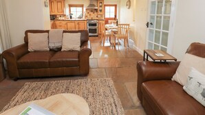 A spacious living area with a brown leather sofa, a wooden coffee table, and a dining table with chairs. A kitchen with wooden cabinets and a stove is visible in the background. - Nick Jone (Richmond)