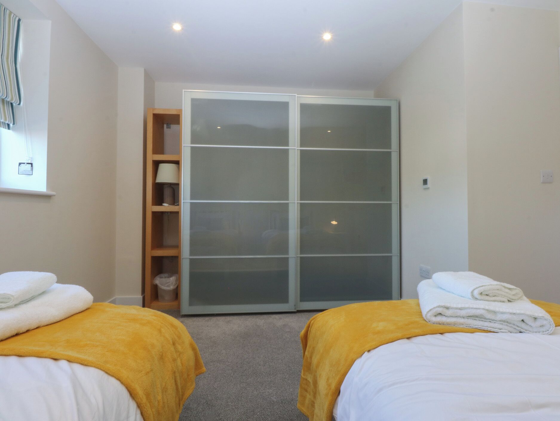 A hotel room with two beds, a wardrobe, a shelf with a lamp, and a window with curtains.