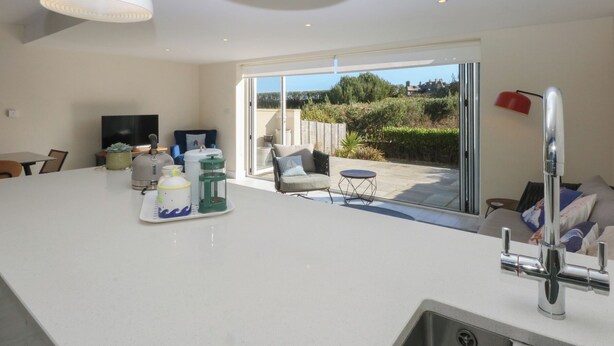 A modern kitchen with a large island, a dining area, and a sliding glass door leading to a patio with a view of the countryside. - 2 Lon Y Dryw (Holyhead)