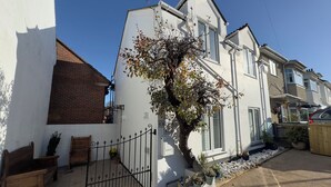 A white two-story house with a brick section on the left, a small garden with a tree, and a wooden bench. - Bohemian Beach Cottage (Bournemouth)