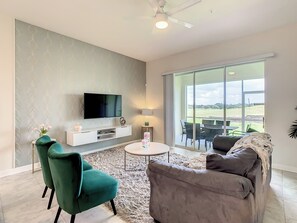 A living room with a flat-screen TV, a green armchair, a grey sofa, a small round table, and a view of the outdoors through large sliding glass doors. - Modern Champions Gate Home Near Parks 2 Beds/2 Baths/ 6 People/Game Room (KISSIMMEE)