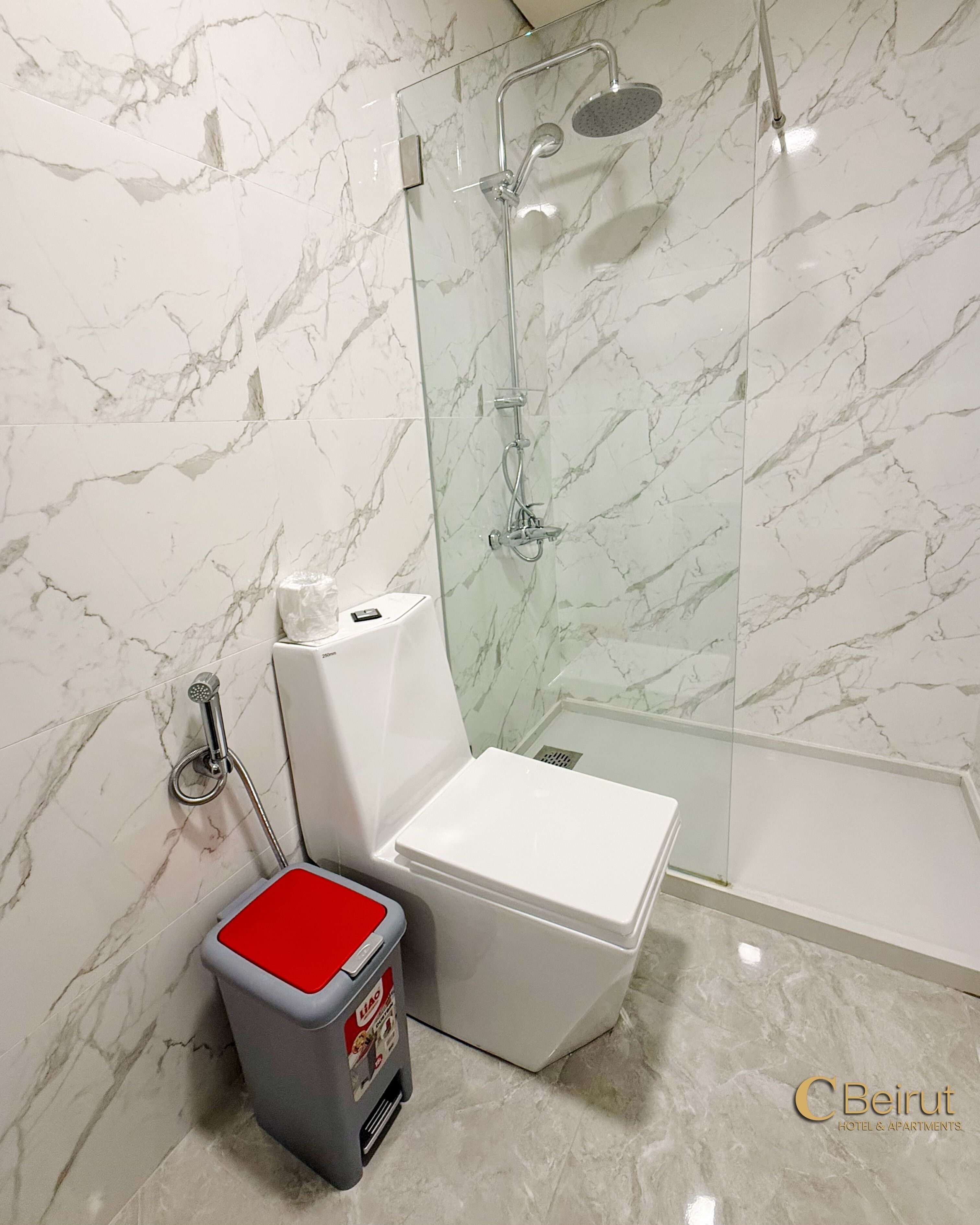 A bathroom with a glass-enclosed shower, a white toilet, and a trash bin with a red top.