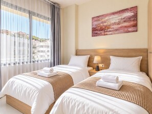 Habitación de hotel con dos camas, un ventanal con cortinas, un cuadro en la pared y una mesita de noche con una lámpara.