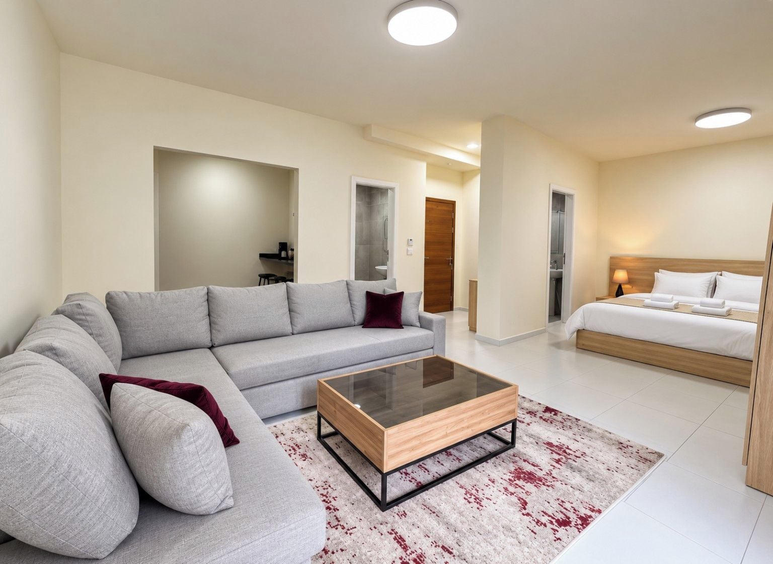 Een moderne hotelkamer met een grijze bank, een houten bijzettafeltje, een bed met witte lakens en een tapijt met een rood-wit patroon.