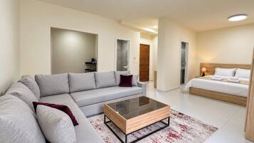 A modern hotel room with a grey sofa, a wooden coffee table, a bed with white linens, and a red and white patterned rug.