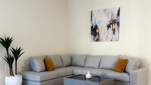 Una sala de estar moderna con un sofá seccional gris, una mesa de centro de vidrio, una planta en maceta blanca y un cuadro abstracto grande en la pared.