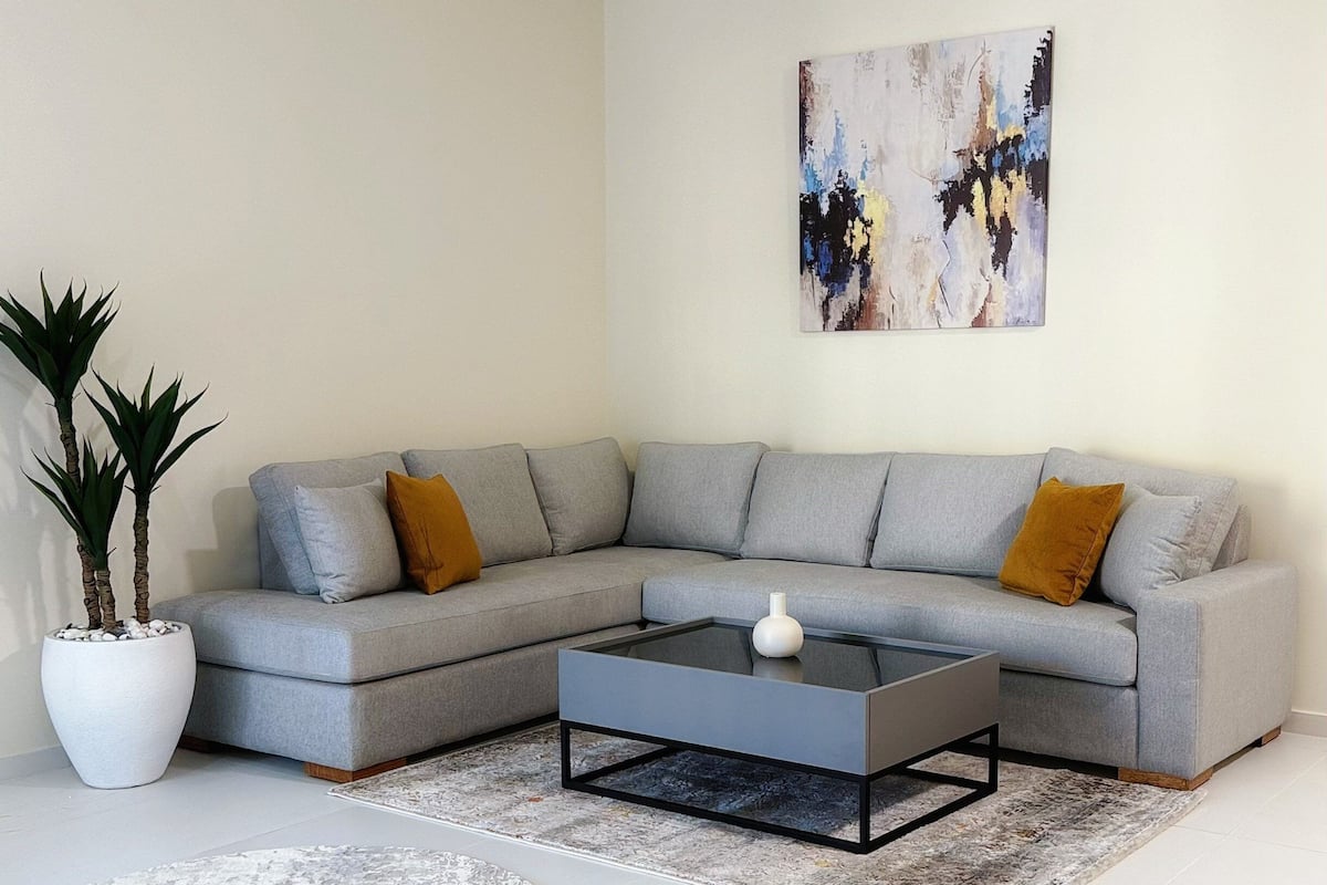 Una sala de estar moderna con un sofá seccional gris, una mesa de centro de vidrio, una planta en maceta blanca y un cuadro abstracto grande en la pared.