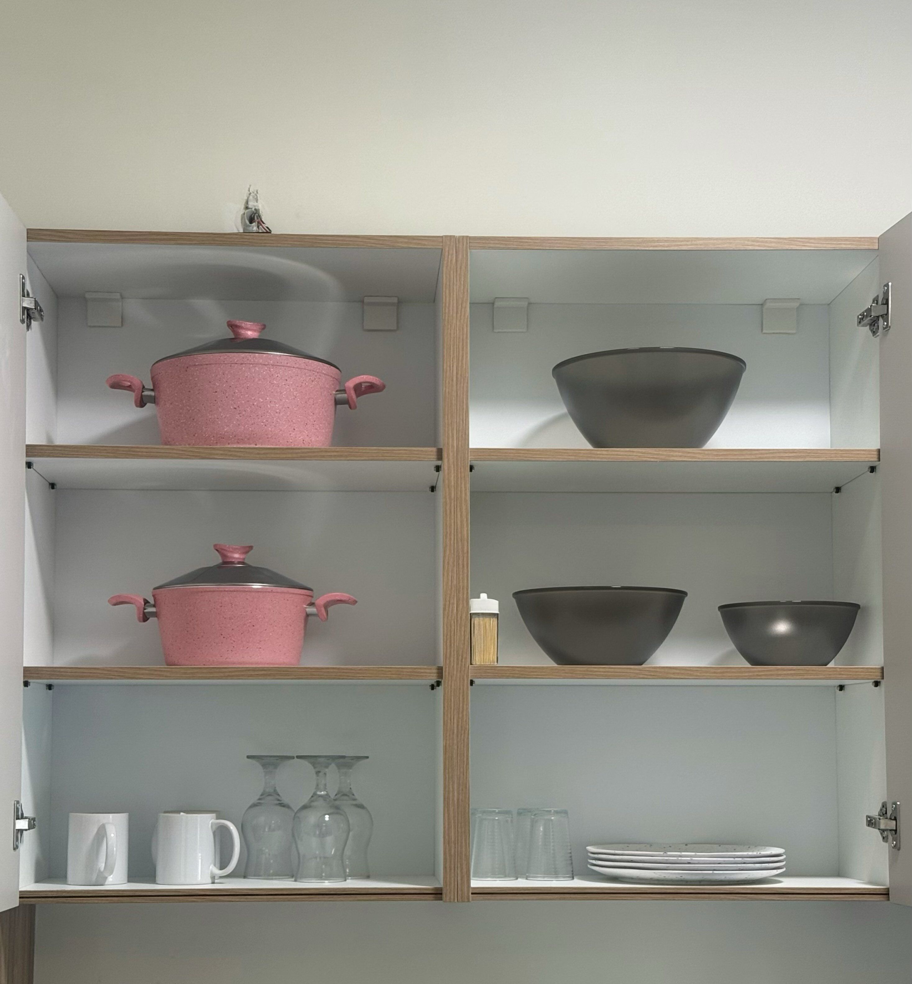 A kitchen cabinet with shelves containing a pink pot, two bowls, a stack of plates, and a few glasses.