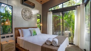 Un dormitorio con una cama grande, una mesita de noche, aire acondicionado de pared y vistas al verdor por la ventana.