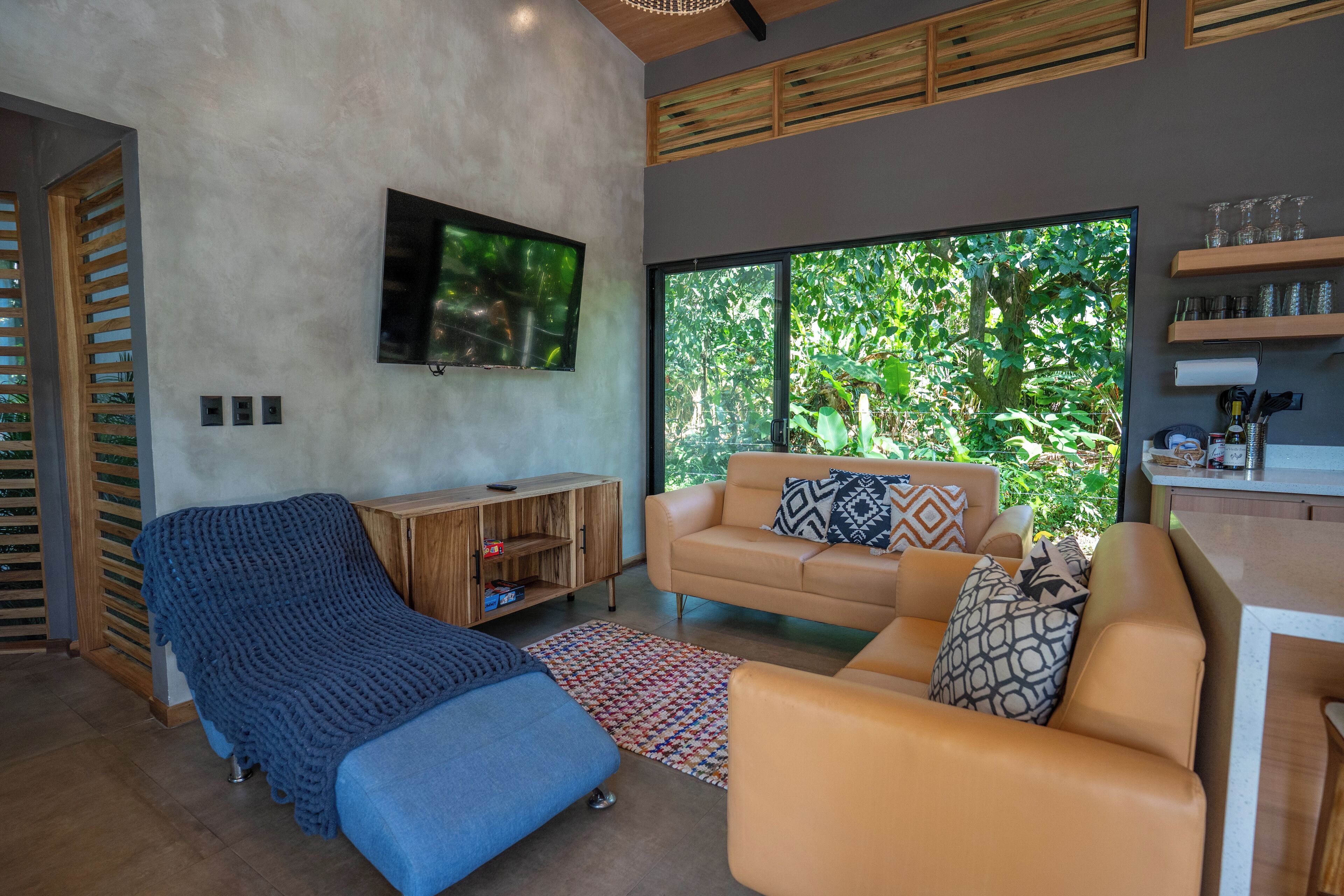 Un salon moderne avec un canapé, un fauteuil bleu, un téléviseur fixé au mur et une grande fenêtre donnant sur un paysage verdoyant.