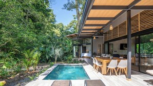 Un área de piscina moderna al aire libre con un deck de madera, sillones de descanso y una mesa de comedor con sillas.