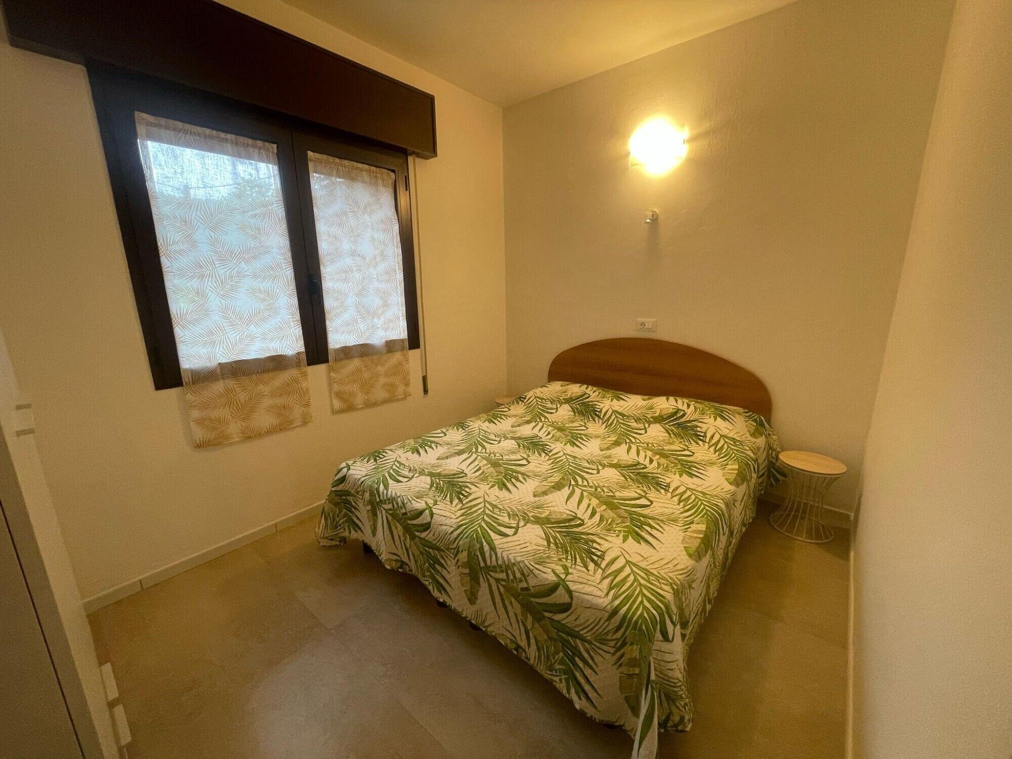 A small bedroom with a single bed, a nightstand, a wall-mounted light, and a window with curtains.