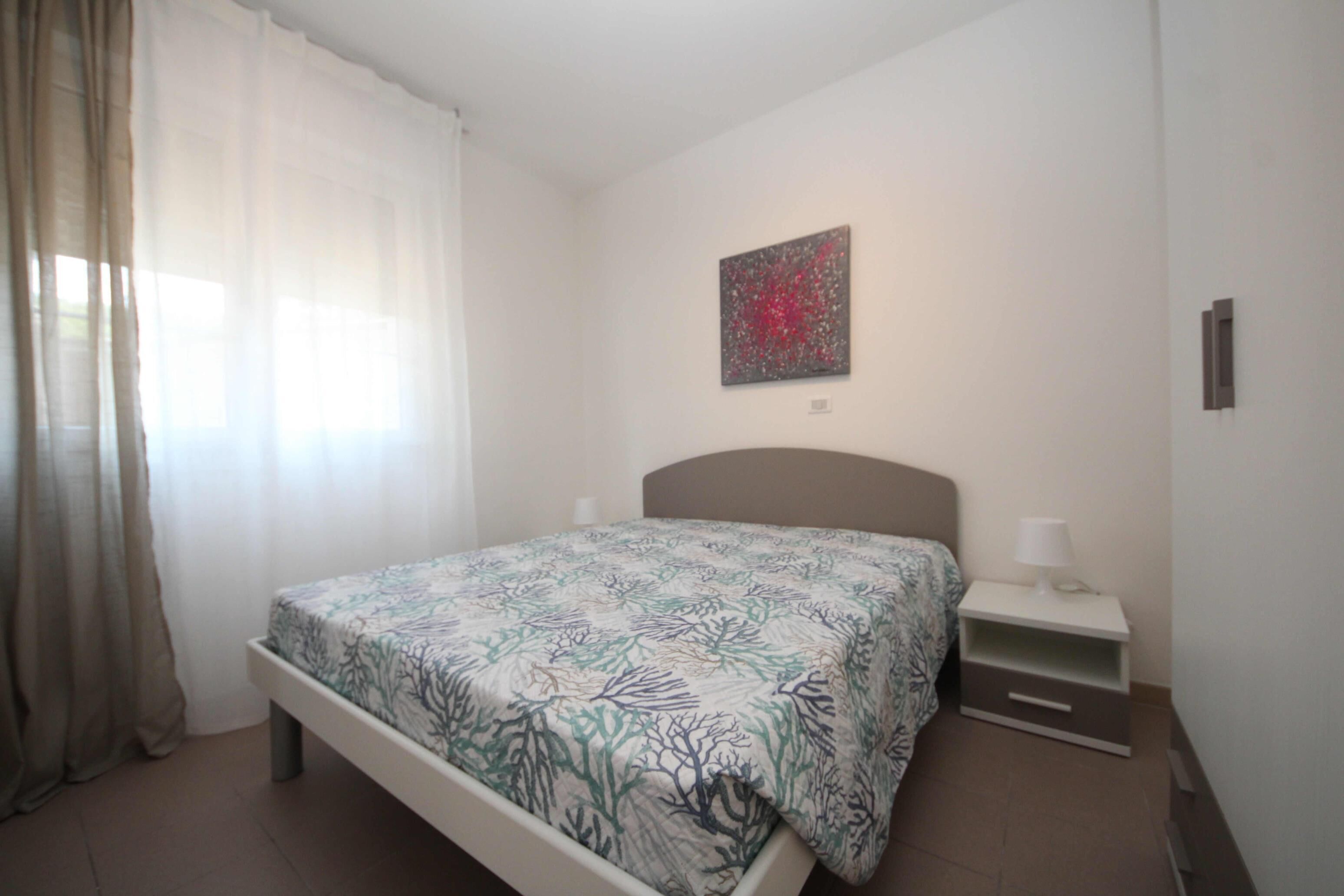 A bedroom with a bed, a nightstand, a lamp, a painting on the wall, and a window with curtains.