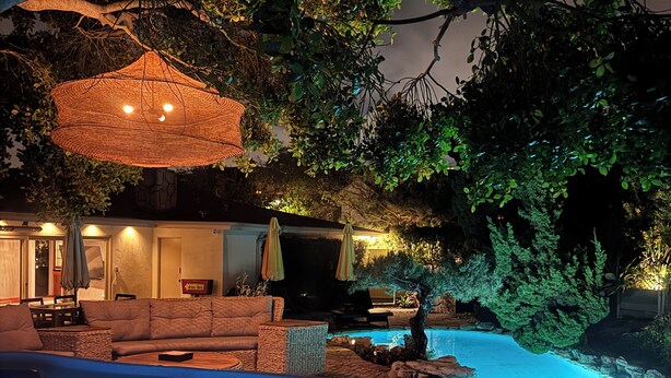 A nighttime outdoor pool area with a hot tub, lounge chairs, and a dining area under a large tree. - Bel Air Mid-Mod Oasis w/ Grotto Pool + Jacuzzi (Los Angeles)