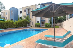 A swimming pool with lounge chairs and an umbrella, surrounded by multi-story buildings with balconies. - Libi Apartment-BlueWaters Property  (Accra)