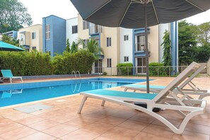 A swimming pool with lounge chairs and an umbrella, surrounded by apartment buildings. - Libi Apartment-BlueWaters Property  (Accra)
