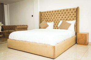 A large bed with a tufted headboard, a bedside table, and a sofa in a modern room with a tiled floor. - Libi Apartment-BlueWaters Property  (Accra)