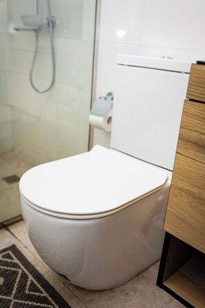 A modern bathroom with a white toilet, a shower area, and a wooden cabinet. - Libi Apartment-BlueWaters Property  (Accra)
