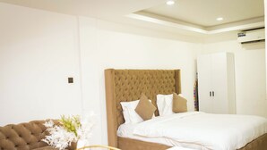A bedroom with a tufted headboard, a large bed with white bedding, a brown sofa, and a small round table with a vase of flowers. - Libi Apartment-BlueWaters Property  (Accra)