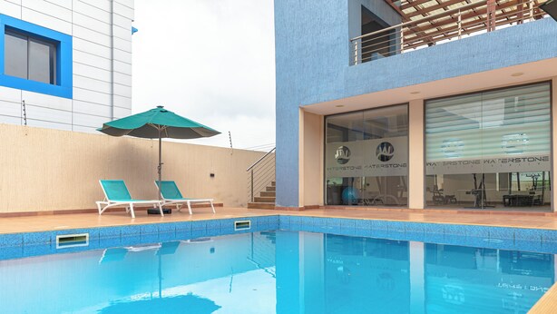 A swimming pool with two lounge chairs and a green umbrella, adjacent to a building with a blue window frame and a glass door with 'Waterstone' signage. - Libi Apartment-BlueWaters Property  (Accra)