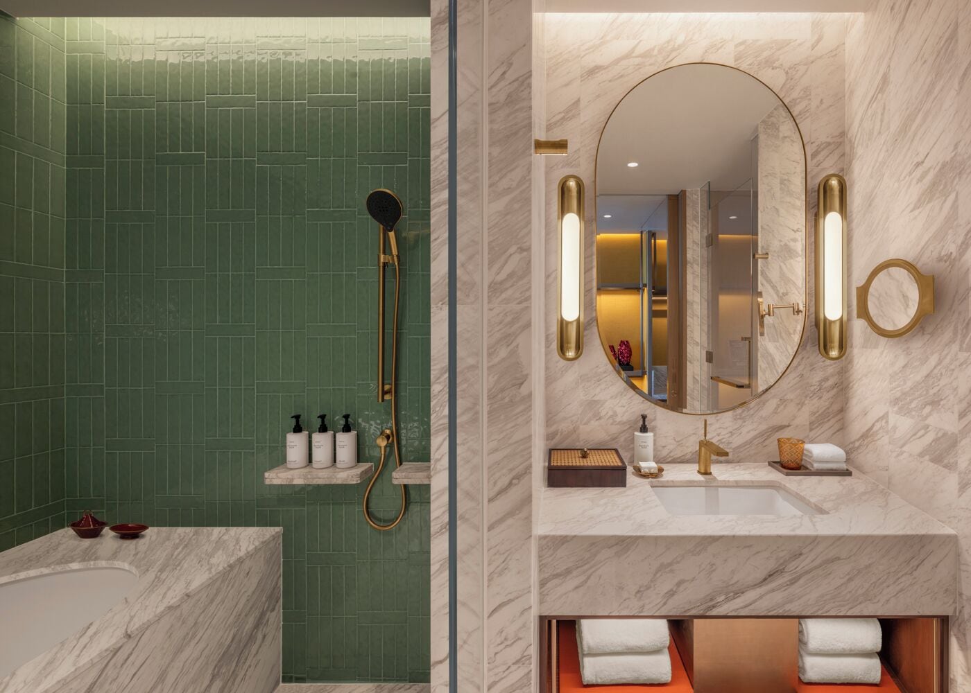 A bathroom with a green tiled wall, a gold showerhead, and a marble sink with a large mirror and wall sconces.