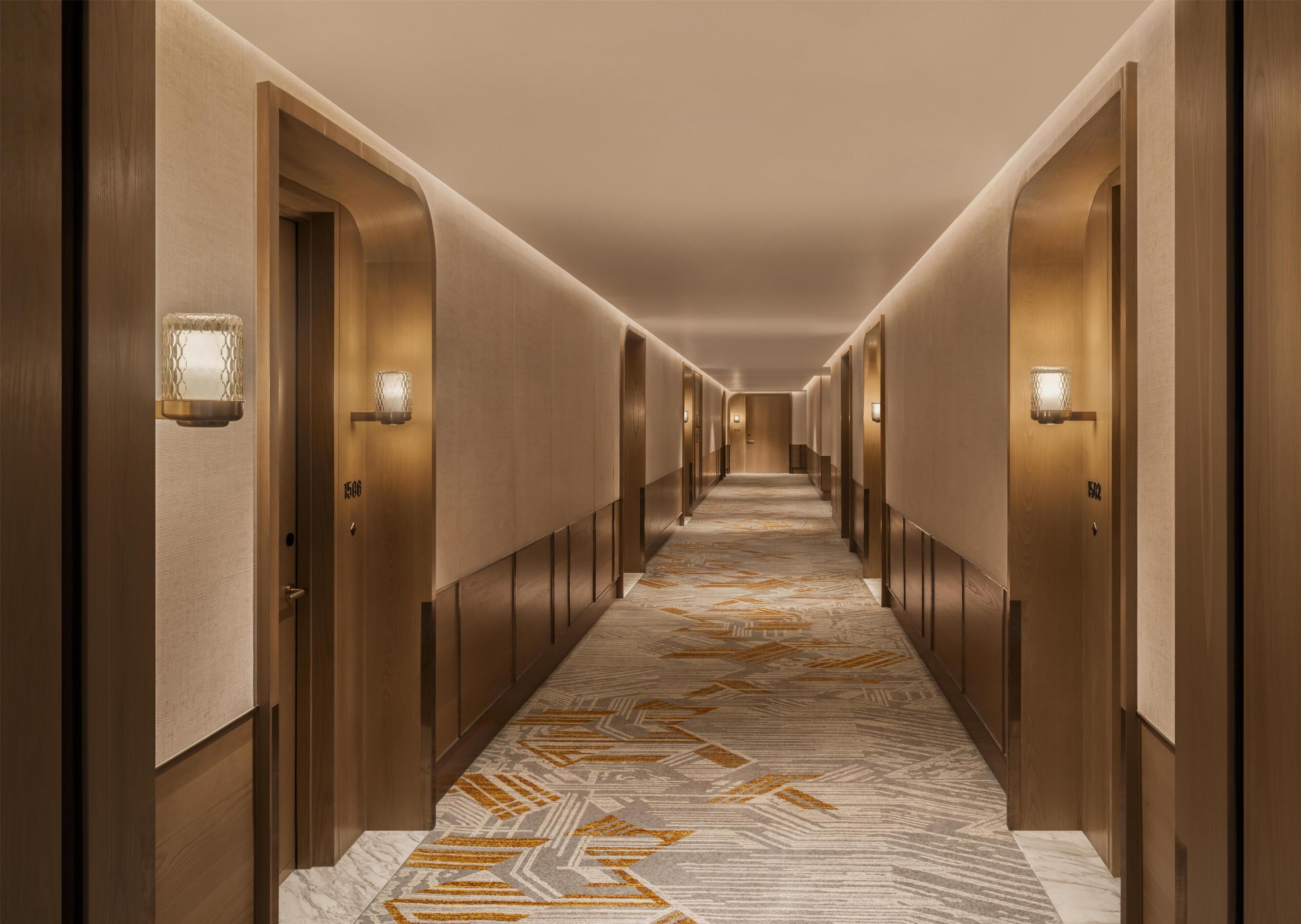 A long, narrow hotel corridor with wooden doors, numbered room signs, and patterned carpet.