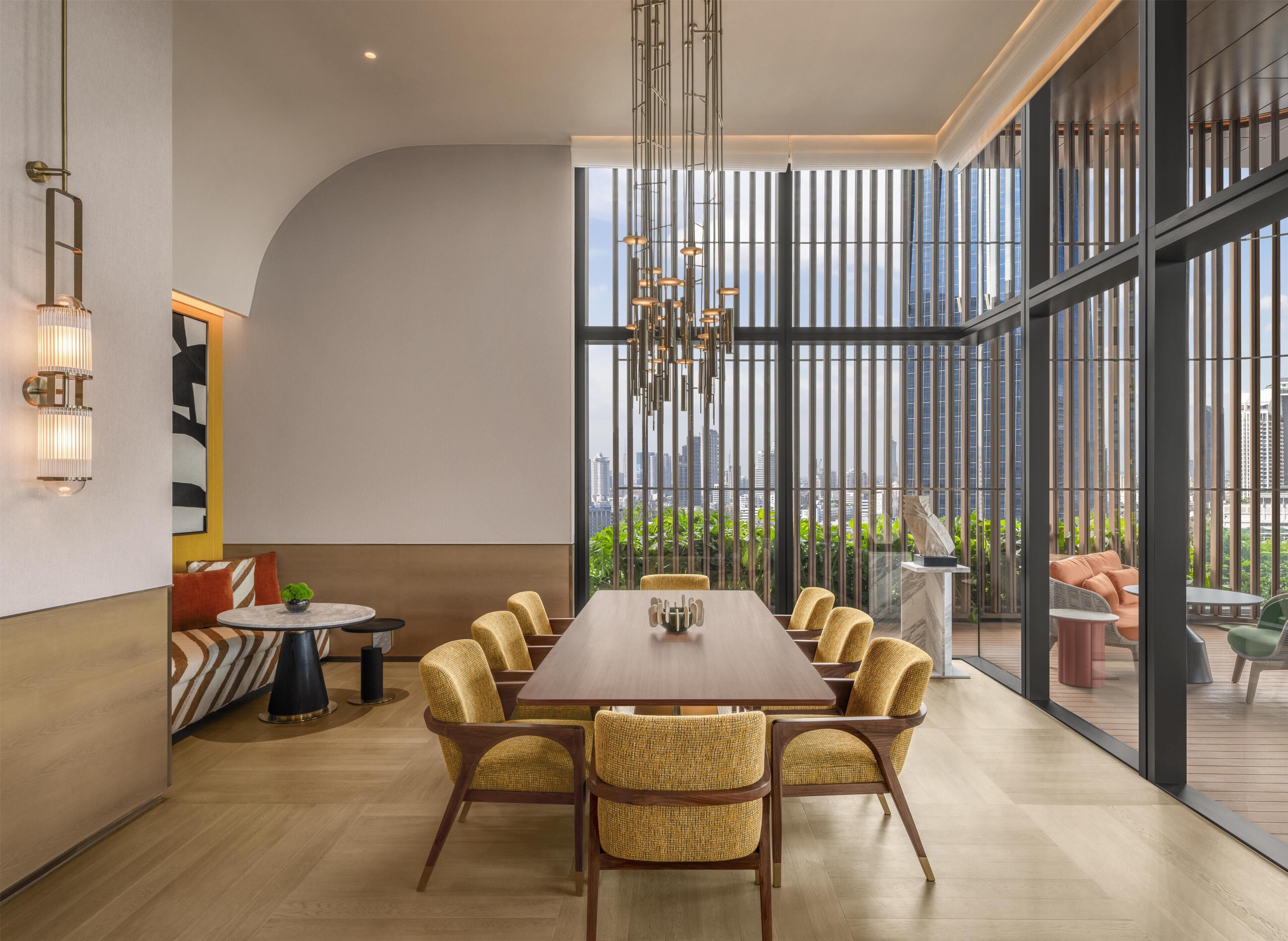 A modern dining area with a long table and wicker chairs, large windows with a city view, and a ceiling light fixture.