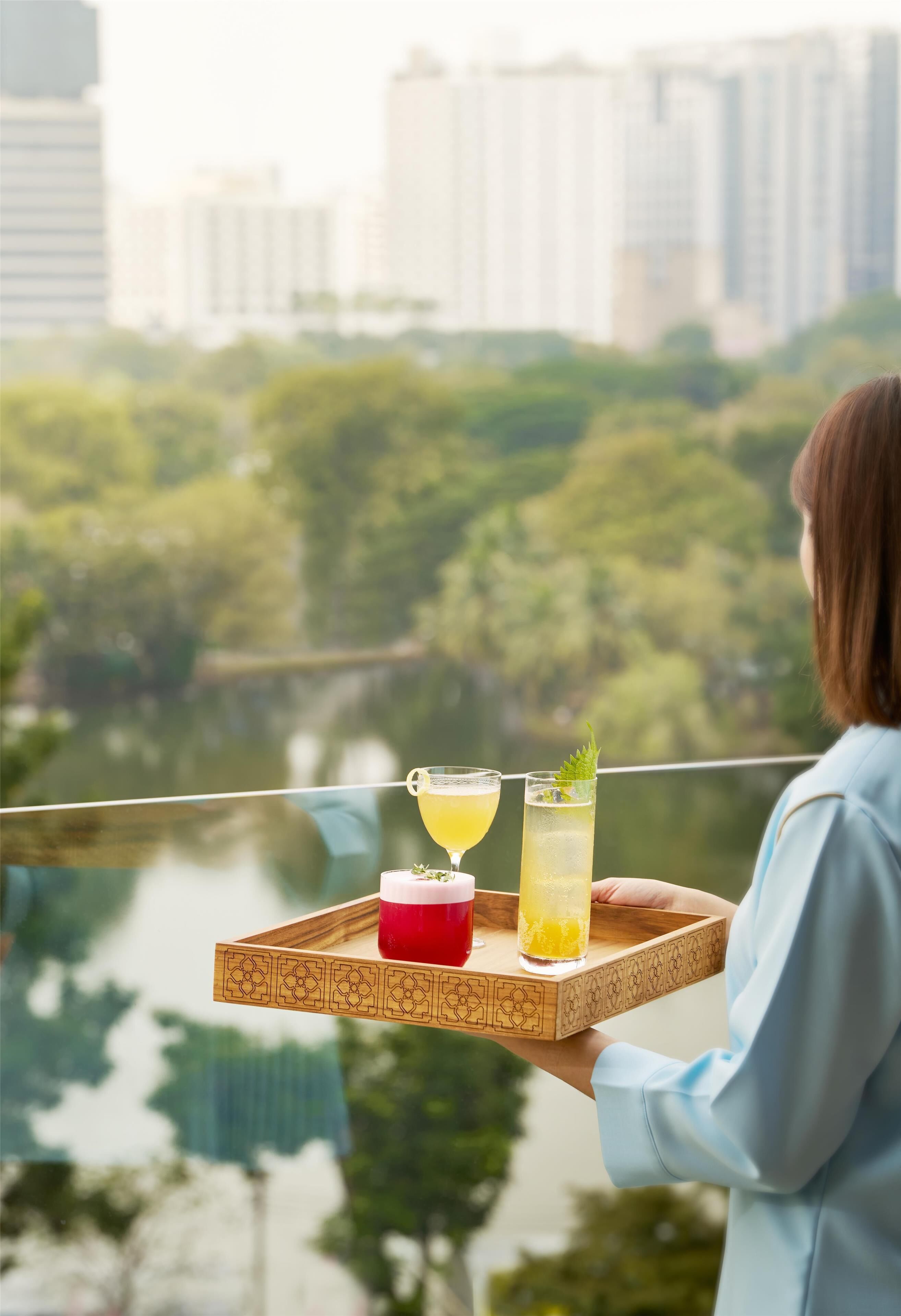Una persona sostiene una bandeja con dos bebidas y un postre, con vista a un río y follaje.