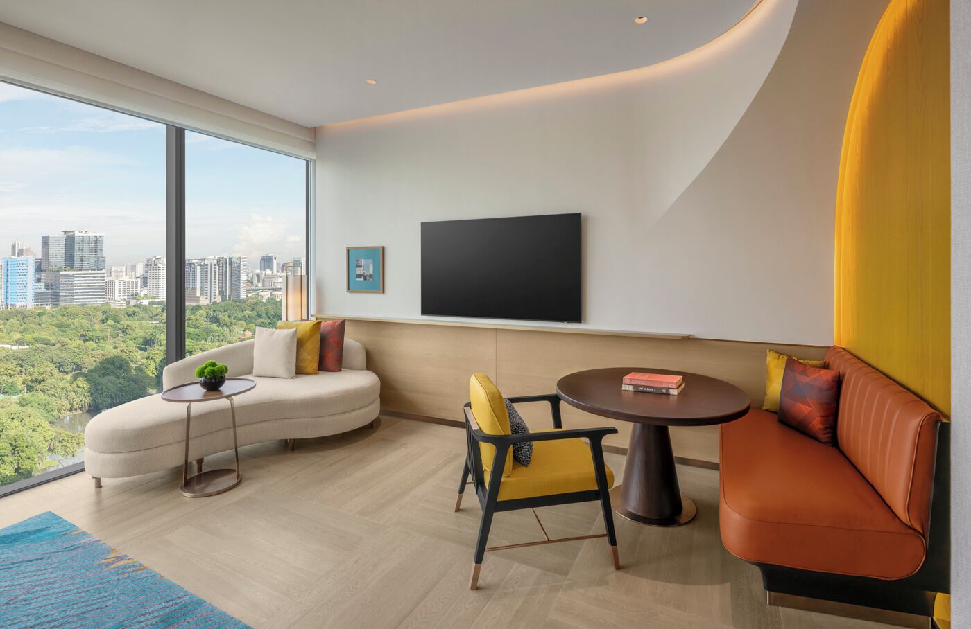 A modern living room with a curved sofa, a small round table, and a chair. A flat-screen TV is mounted on the wall, and a city view is visible through large windows.
