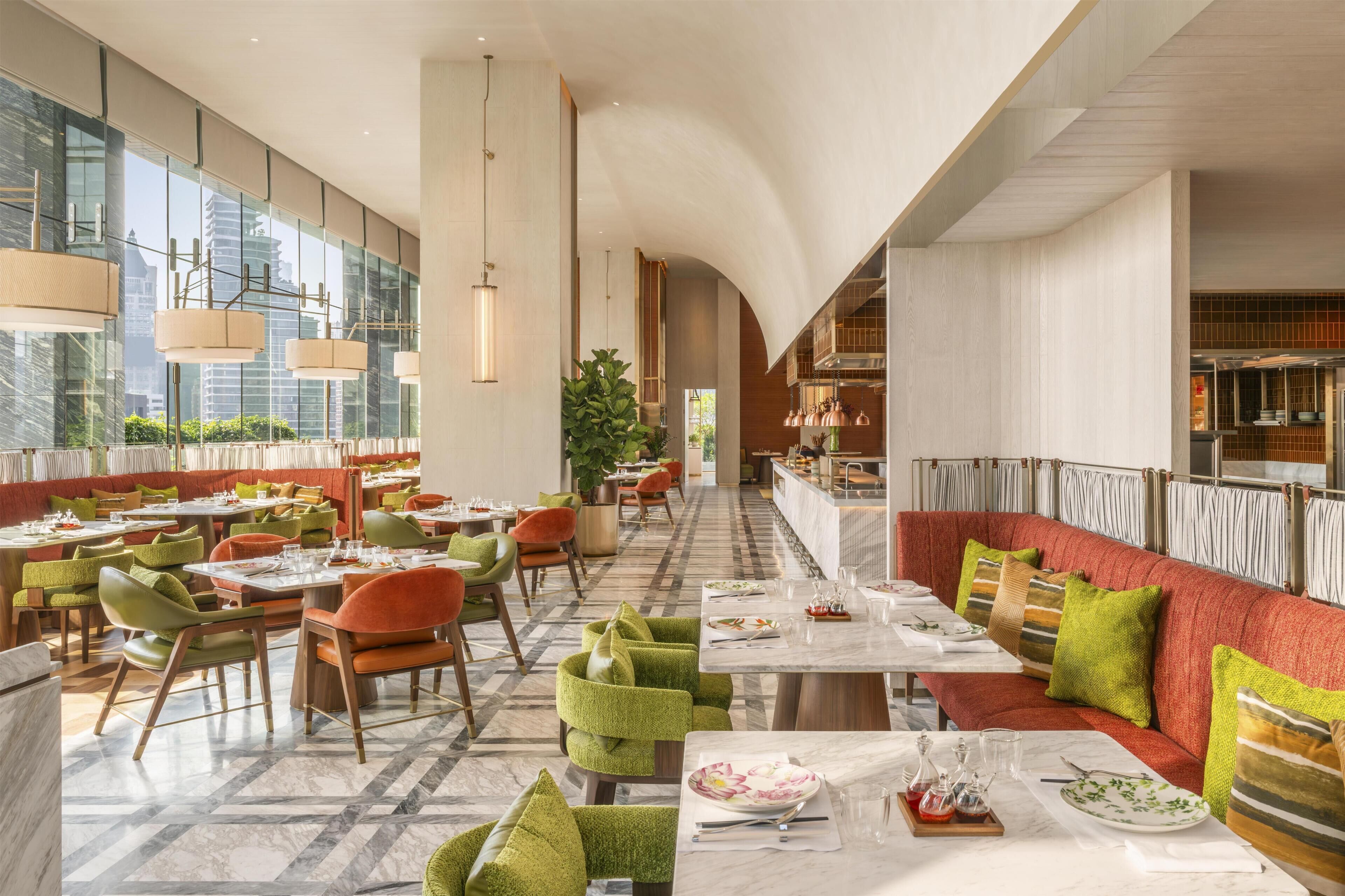 A modern restaurant with high ceilings, large windows, and a mix of seating options including tables and a long red bench.