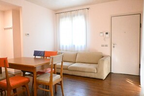 Ein Zimmer mit einem Holzesstisch, zwei Stühlen, einem beigen Sofa, einer weißen Tür und einem Fenster mit durchsichtigen Vorhängen.