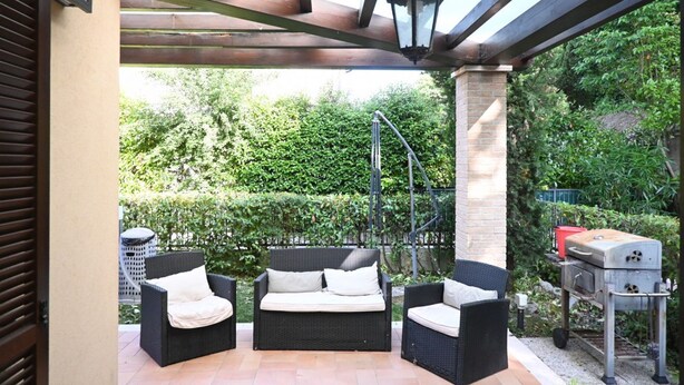 Een terras buiten met rieten meubels, een barbecue en een pergola.