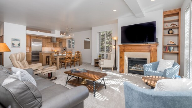 A modern living room with a grey sofa, a blue armchair, and a coffee table. There's a fireplace, a TV mounted on the wall, and a kitchen in the background. - Walk to Skiing, Dining and Downtown (Ketchum)