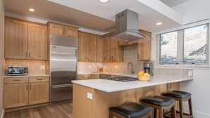 A modern kitchen with wooden cabinets, a stainless steel refrigerator, a built-in oven, and a large island with bar stools. - Walk to Skiing, Dining and Downtown (Ketchum)