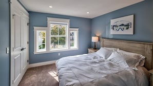 Ein Schlafzimmer mit einem Bett, einem Nachttisch, einer Lampe, einem Fenster und einem Gemälde an der Wand.