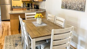 Un comedor con una mesa de madera, sillas blancas, un tapete y una cocina al fondo.