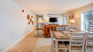 A modern living room with a sofa, dining table, and chairs, a TV, and a window with a view.
