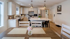A modern kitchen with white cabinets, a central island, pendant lighting, and a dining area with a table and chairs. - Cady Suite at 109 Main (Stowe)