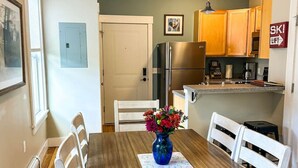A dining area with a wooden table, white chairs, a vase with flowers, and a kitchen in the background. - Pinnacle Suite at 109 Main (Stowe)