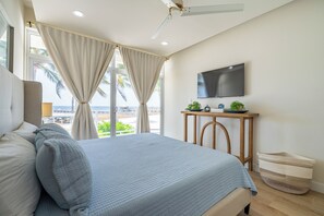A bedroom with a bed, curtains, a television, a small table with plants, and a view of the ocean. - Styles Beach 1A - Beachfront Luxury Condo w/ Pool (San Pedro Town)