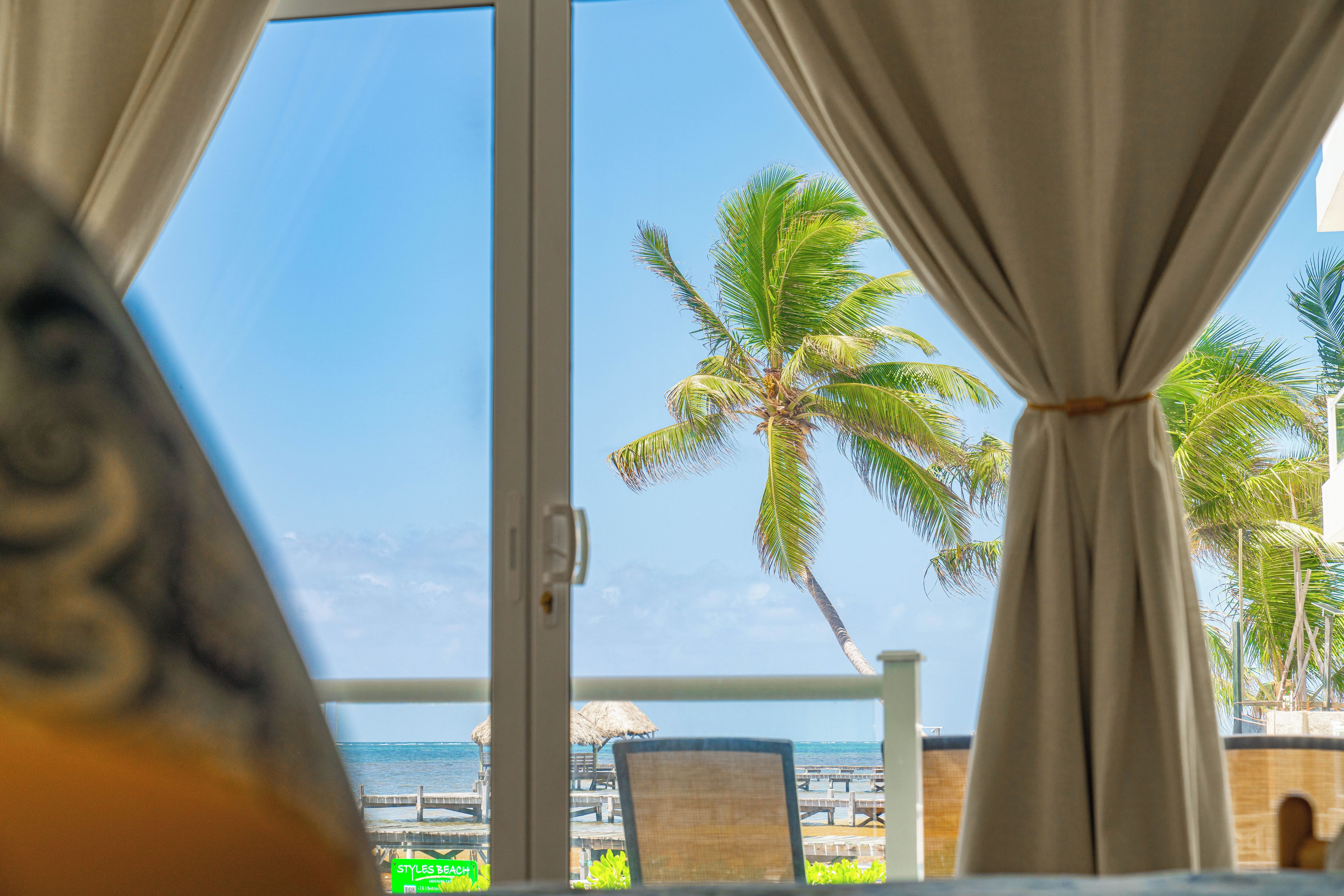 Vista desde una ventana con cortinas, mostrando una palmera y una playa con muelle de madera.