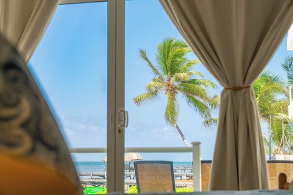 A view from a window with curtains, featuring a palm tree and a beach with a wooden pier. - Styles Beach 1A - Beachfront Luxury Condo w/ Pool (San Pedro Town)