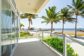 A balcony with a view of the beach, palm trees, and a thatched-roof structure. - Styles Beach 1A - Beachfront Luxury Condo w/ Pool (San Pedro Town)