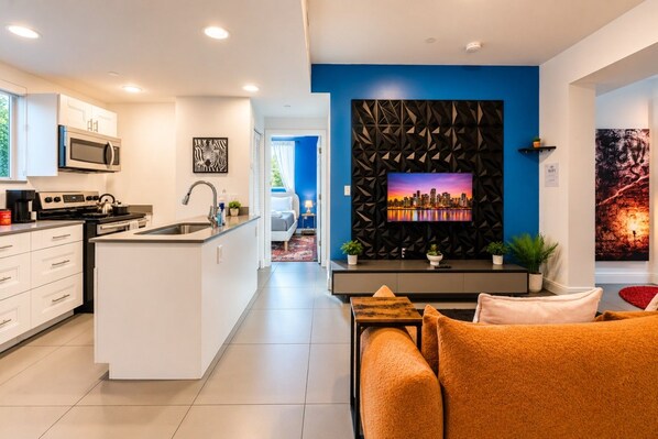 Eine moderne Küche mit integriertem Mikrowellenherd und Herd, ein Essbereich mit Sofa und Couchtisch sowie ein Wohnzimmer mit Fernseher und dekorativer Wandverkleidung.